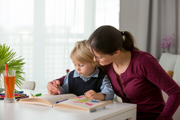 Cute preschool child, blond boy, filling some homework in a work book and coloring, mother helping him
