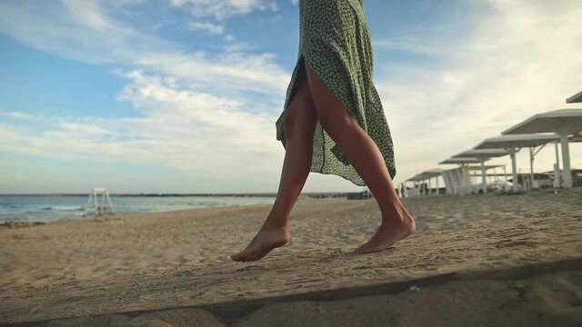 Legs of a young woman in slow motion walking barefoot on the beach. Leaving footprints in the sand Tourist on vacation by the sea. Woman in a beautiful fluttering dress in the wind