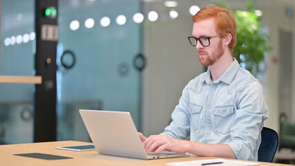 Focused Young Redhead Man Working on Laptop in Office 