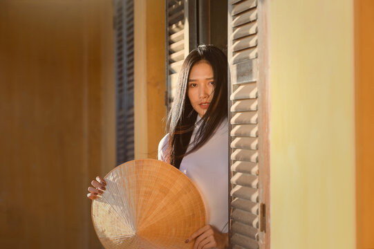 Beautiful woman standing in a doorway holding a traditional non la conical hat, Thailand