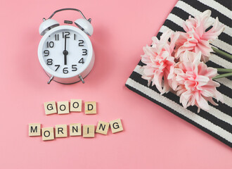 flat lay of white vintage alarm clock  with wooden letter "GOOD MORNING" and bouquet of pink flower on black and white stripes cloth on pink background.