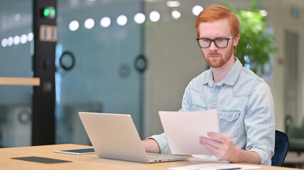 Young Redhead Man Working on Laptop with Documents in Office 