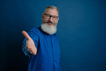 Mature bearded caucasian man holds out his hand to say hello. Friendly grayhaired bearded senior man dressed blue shirt makes greeting gesture over blue background.