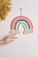 Interior braided decoration made as soft-colored rainbow with cotton tassels is hanging on the wall near craft paper sun. Female hand is touching wall ornament for child or baby room. 