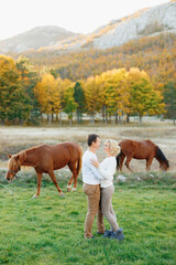 Man hugs woman against the background of grazing horses in the autumn forest