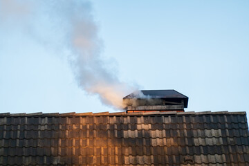 A fragment of the roof of a house covered with black ceramic tiles. From the chimney comes the smoke from the stove. Background.