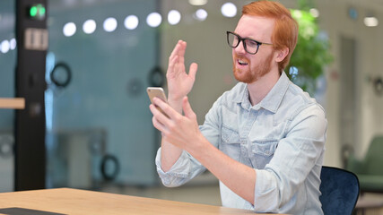 Discouraged Redhead Man Reacting to Loss on Smartphone in Office 