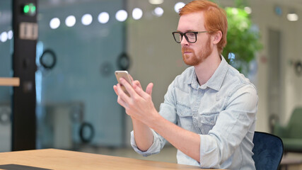Serious Young Redhead Man using Smartphone in Office 