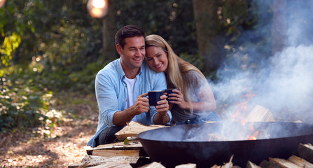 Romantic Couple Camping Sitting By Bonfire In Fire Bowl With Hot Drinks