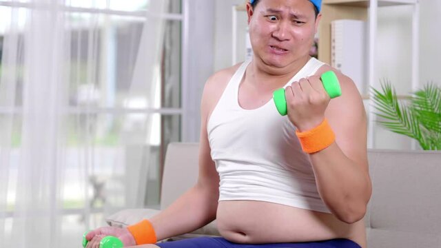 Funny fat Asian guy in sportswear lifting two side dumbbell with difficultly in living room. He intend harder to weight training bicep on couch and show belly. Chubby man tired to lift by both hands