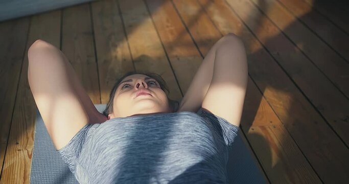Woman in grey t-shirt does crunches on grey mat on parquet floor in room with sunbeams and shadows close view slow motion