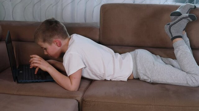Thoughtful boy in white shirt lying on comfy sofa at home studying on laptop computer . Serious schoolboy playing video games at home.