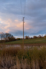 Electric pole with wires at the ditch with bushes on the grass, cultivated land and trees. Blue...