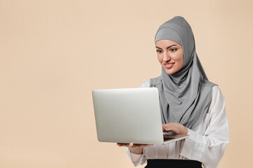 Smart Middle Eastern muslim islamic arabian student female woman using laptop for online learning, webinars isolated over beige background. Clever office worker girl in hijab using laptop