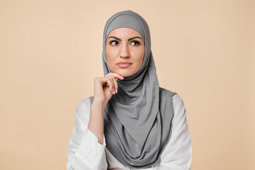 Thoughtful young arabian middle eastern muslim islamic woman thinking about shopping, love, relationship isolated on beige background. Pensive woman in grey hijab making plans for future