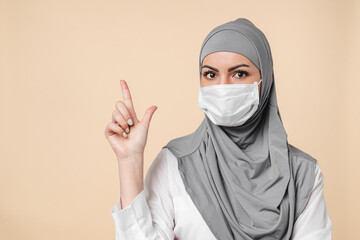 Young Middle eastern islamic muslim arabian woman in protective face mask against Covid 19 coronavirus and grey hijab pointing at copy space isolated over beige background. 