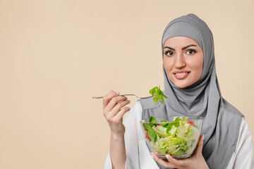 Woman Middle Eastern muslim islamic arabian eating salad from salad bowl in grey hijab isolated on beige background. Young female dieting, healthy eating habits, organic food.