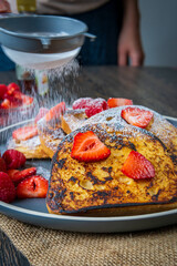 Woman sprinkles sugar powder on a stack of french toasts with fresh strawberries, raspberries and maple syrup. Tasty morning breakfast or brunch with gluten free bread. Easy making and healthy dessert