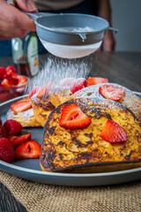 Hand sprinkles sugar powder on a stack of french toasts with fresh strawberries, raspberries and...