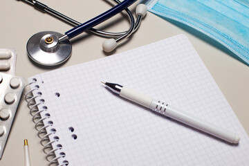 On a light gray background, a medical mask, a stethoscope, pills and a notebook with a pen. Medical concept