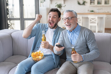 Overexcited energetic football soccer baseball fans old father and adult son rooting cheering for favorite sport team watching match on TV drinking beer on sofa. Happy father`s day! Goal