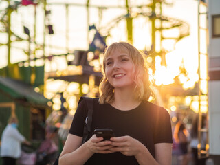 Mujer joven usando su móvil durante la puesta de sol en una feria o parque de atracciones