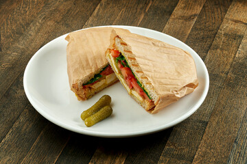 Large fried sandwich with with tofu, tomatoes and pesto sauce, served in parchment on a white plate on a wooden background