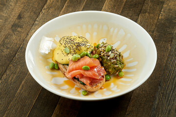 Appetizing breakfast - fried toast with poached egg, salmon and hollandaise sauce garnished with guacamole in a white bowl on a wooden background
