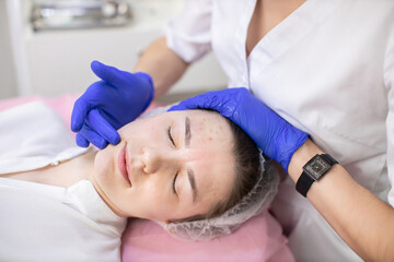 Obraz na płótnie Canvas Young pleasant woman lying on the couch at dermatology center and smiling, while hands of doctor cosmetologist in rubber gloves touching her cheeks while applying lotion or serum