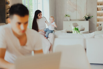 Obraz premium Young mother looks after her baby while her husband works on laptop