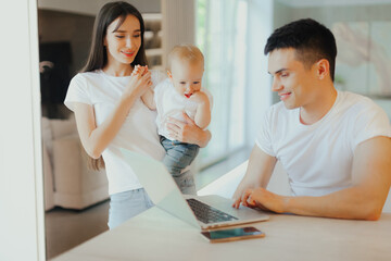 Young father works on laptop while his wife looks after child