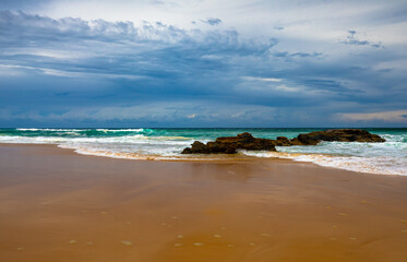 A view of a national park beach with smooth sand, beautiful turquoise water and white surf action framed by the dark clouds of a recent storm, located on the east coast of Australia.