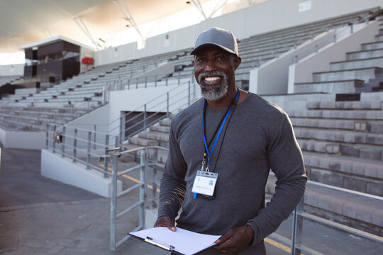Portrait of african american male coach holding clipboard smiling while standing in the stadium
