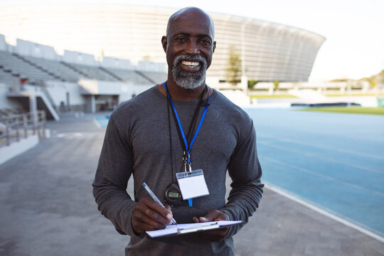 Portrait of african american male coach holding clipboard smiling while standing in the stadium