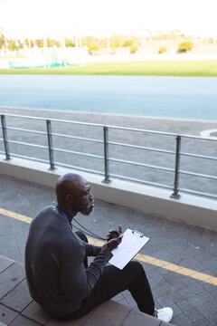 African american male coach with stopwatch measuring time while sitting on the seats in the stadium
