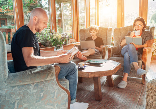 Father, mother, and son family sitting together in Sunroom in cozy armchairs and reading books, using a laptop, and enjoying togetherness. Pandemic lockdown, home office and stay-at-home concept image