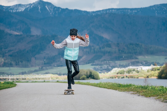 Cute girl skateboarding on the longboard on the asphalt road with a mountain landscape background. She gracefully pushing off and slow riding. Happy outdoor childhood concept image.