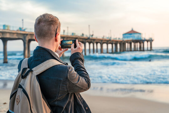 A young man with a backpack takes photos of the pier with his mobile phone on the beach in Los Angeles with a bright sunset