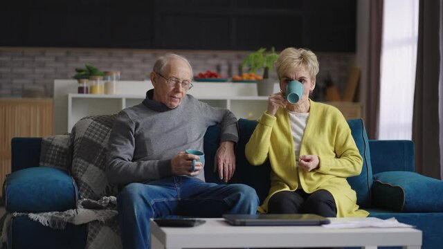 calm married couple of old people is resting at home at weekend, granny and grandpa are drinking tea in living room of apartment