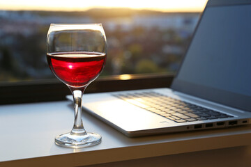 Glass of red wine on background of laptop and window with sunset view. Alcohol for inspiration, relax after work