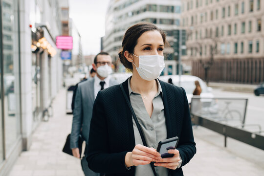 Businesswoman with smart phone standing on footpath in city during COVID-19