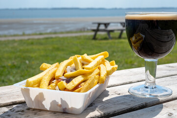 Glass of dark strong belgian beer and portion of fried potato chips with sauce and onion, served on outdoor terrace with green grass meadow on background