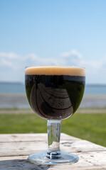 Glass of dark strong belgian beer served on outdoor terrace with green grass meadow on background
