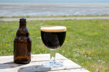 Glass of dark strong belgian beer served on outdoor terrace with green grass meadow on background