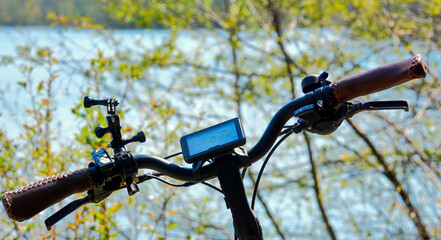 Handlebars of a pedelec with attachments for an action cam in front of the blurred background of a blue water with green shore stripe