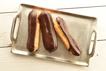 Three chocolate eclairs on a metal tray, close-up, on a wooden table, top view.