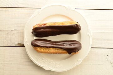 Two chocolate eclairs on a white ceramic plate, close-up, on a wooden table, top view.
