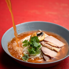 Ramen soup is poured with broth. Bowl of noodles, meat and herbs parsley on red table. Spicy oriental dish, Pan-Asian cuisine.