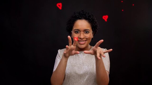 leisure and people concept - african american woman in glasses dancing and drawing heart in air with fingers over black background with animated glowing neon lines