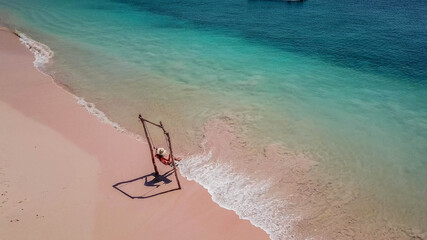 Fototapeta na wymiar A girl swinging on the beach swing on Pink Beach, Lombok, Indonesia. Captured from above with a drone. The water changes colors from turquoise to navy blue. beach has a nice coral color. 